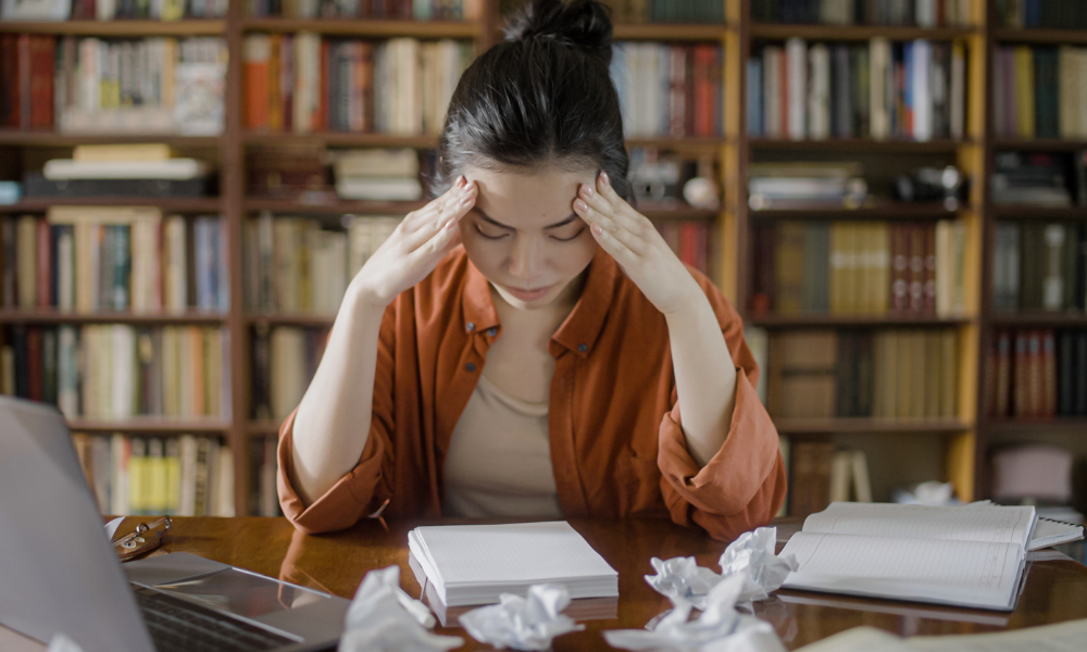 A young woman sits at a desk in a library, holding her head in her hands