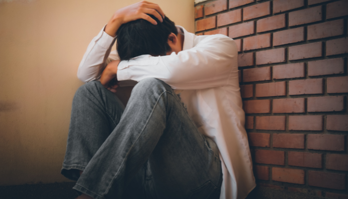 A man is sitting against a wall, head down, showing deep emotional distress or sadness.