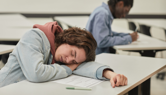 A student asleep in class with another student working in the background.