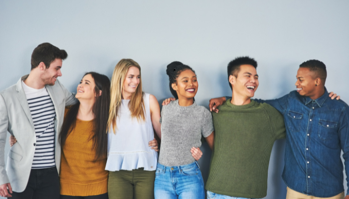 Group of six diverse young people smiling, standing side by side with their arms around each other.